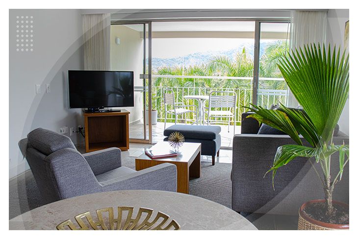 Sala moderna con muebles grises, televisor, mesa de centro y acceso a un balcón con sillas y una vista llena de palmeras que resalta la experiencia en los mejores hoteles de Ibagué