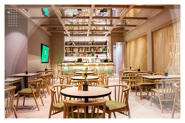 Restaurante moderno con sillas de madera clara, mesas redondas, iluminación cálida y una barra al fondo decorada con botellas y plantas que reflejan el ambiente acogedor del hotel Estelar Ibagué