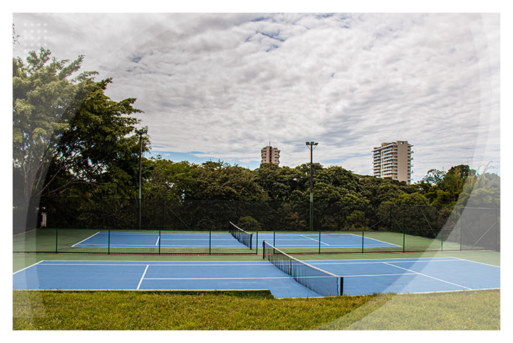 Dos canchas de tenis azules al aire libre rodeadas de árboles y edificios al fondo, perfectas para disfrutar del deporte con la promoción de hoteles todo incluido en Ibagué