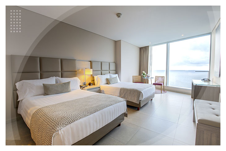 Habitación doble con diseño moderno, camas con sábanas blancas, ventanal de piso a techo con vista al mar y muebles en tonos claros, ideal para quienes buscan hoteles cerca al mar en Cartagena