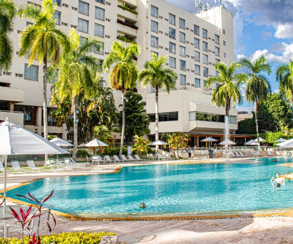 Piscina de agua cristalina, ubicada al aire libre en un hotel que está rodeado de grandes palmeras y un edificio de color blanco con varios pisos al fondo, a la que se acceder con los paquetes de Hoteles Estelar con descuento