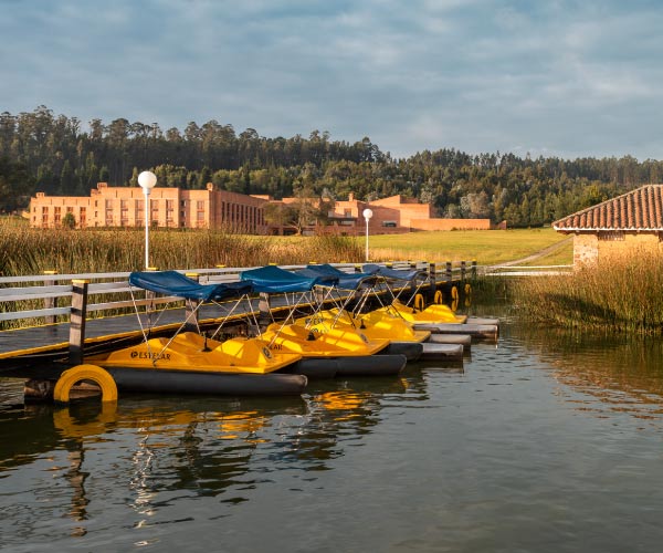 Muelle que tiene bicicletas acuáticas de color amarillo con toldo azul, incluidas con el paquete de viajes con descuento en Hoteles Estelar, rodeado de un entorno con vegetación y un edificio de ladrillo