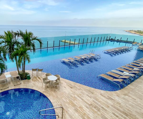 Piscina infinita con palmeras, que tiene vista al mar y una zona de descanso con sillas asoleadoras dentro del agua, que se pueden disfrutar al hacer reservas de Hoteles Estelar con descuento en Colombia