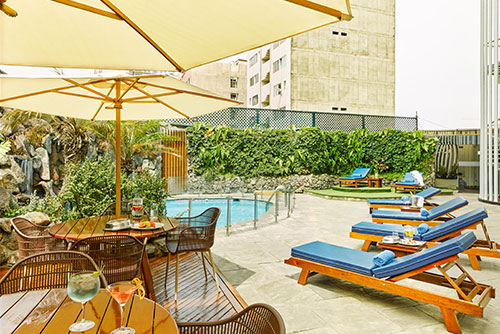Fotografía en la que se ven mesas de madera, sillas asoleadoras con cojín azul, una piscina ubicada en el centro del lugar; todo esto rodeado de edificios, vegetación e incluido en el paquete de viaje a Perú de hotel Estelar y Compensar