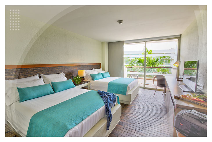Habitación amplia con dos camas, cobijas color azul, escritorio, televisión, y balcón con vista a las palmeras, perfecta para descansar en el Hotel Estelar Playa Manzanillo Cartagena