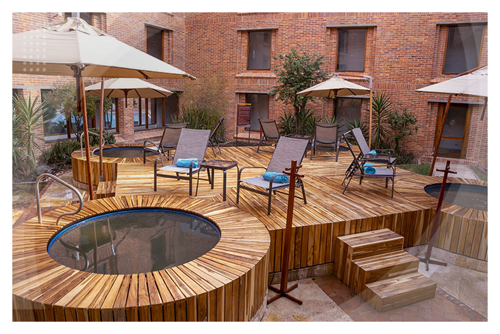 Zona de descanso en terraza de madera con sillas, sombrillas y pequeños jacuzzis al aire libre rodeados de un edificio de ladrillo en un hotel cerca al lago Sochagota
