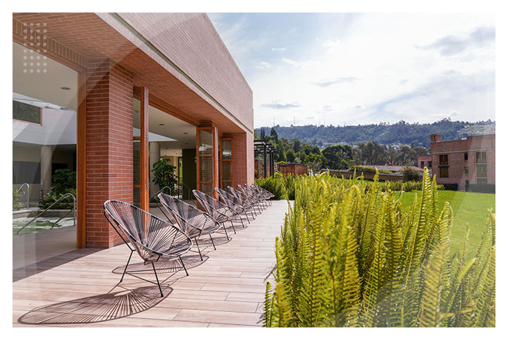 Terraza exterior con sillas metálicas junto a un edificio de ladrillo rodeado de plantas verdes y montañas al fondo en uno de los hoteles en Paipa