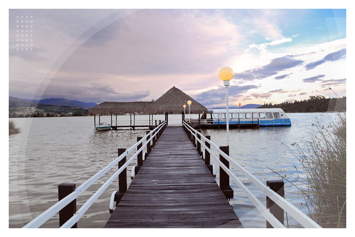 Muelle de madera con barandas blancas que conduce a una estructura con techo de palma sobre el agua al atardecer, con montañas al fondo y una lancha como parte de la oferta de hoteles en Paipa
