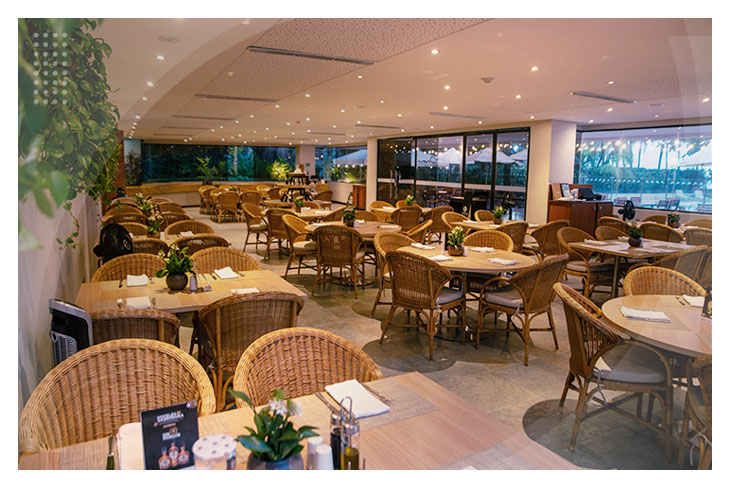 Espacioso restaurante con múltiples mesas y sillas de mimbre, rodeado de ventanales que permiten ver zonas verdes, como parte de los servicios de los mejores hoteles en Santa Marta