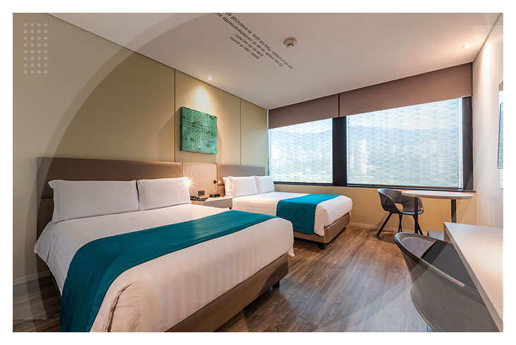 Dos camas dobles con sábanas blancas y una manta decorativa de color azul turquesa de la habitación por una noche en el Hotel Estelar Square, que cuenta con un escritorio y un ventanal con vista a la ciudad