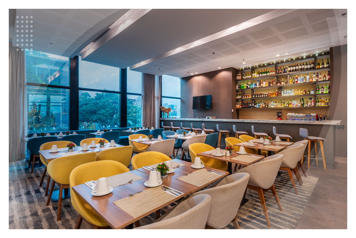 Restaurante moderno con sillas de colores amarillo y gris, que hace parte de los paquetes de viaje con el Hotel Estelar Square, acompañado de un gran ventanal en donde se puede contemplar la naturaleza