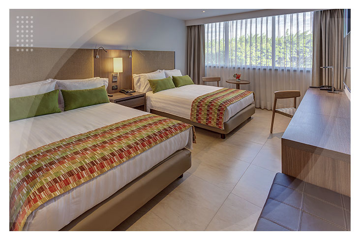 Habitación con dos camas amplias, cojines verdes, escritorio de madera y una gran ventana con cortinas que permiten la entrada de la luz natural en el Hotel Estelar Villavicencio