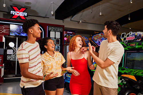 Dos hombres y dos mujeres, que visten ropa casual para clima cálido de colores blanco, negro, rojo, amarillo, beige y café claro, están de pie sonriendo y divirtiéndose en el área de juegos de Lagomar en la cual hay diferentes máquinas recreativas