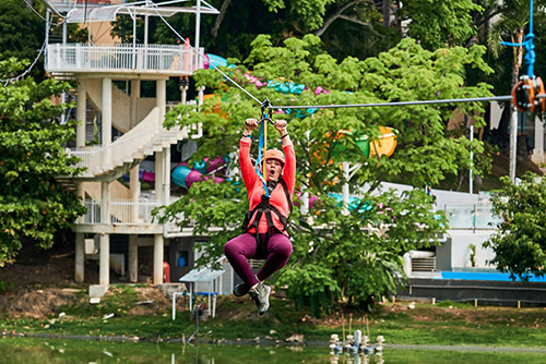 Una mujer, que viste una camisa de manga larga coral, leggins morados, arnés y casco de protección, extiende sus manos hacia arriba para sujetarse de una polea y bajar por el cable de la tirolesa de Lagomar, que la desliza sobre un Lago