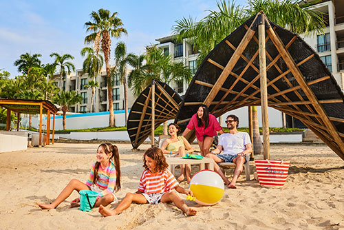 Dos mujeres, un hombre, un niño y una niña, que visten ropas de clima cálido de colores fucsia, blanco, amarillo, naranja, rosado y azul, se encuentran sentados junto a un parasol, una mesa, un bolso y una pelota de playa, disfrutando de la diversión y relajación de los planes turísticos en Compensar