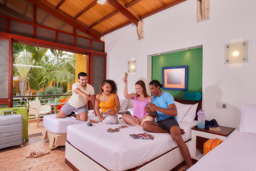 Cuatro personas están compartiendo juntos en una habitación que tiene tres camas de sabanas de color blanco, que están incluidas al hacer una reserva de habitación en hotel Lagosol. En el fondo se puede ver una ventana, la cual deja ver la vegetación que esta en el exterior​