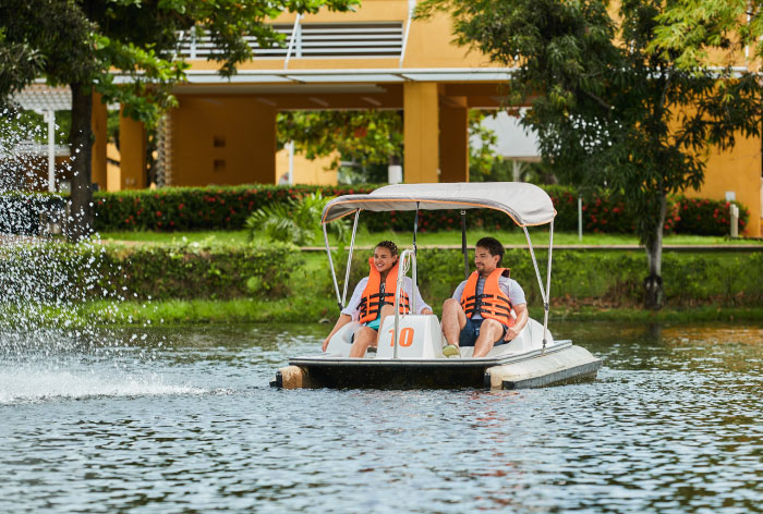 Pareja de personas jóvenes que usan chaleco salvavidas anaranjado están navegando en un bote de color blanco, en medio de un lago de las instalaciones del hotel Lagosol de Compensar. Alrededor se pueden ver edificios de color mostaza y vegetación