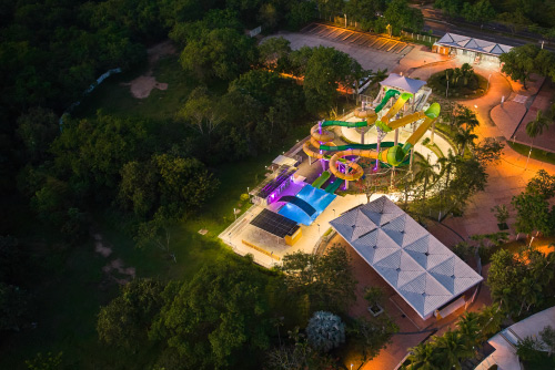 Toma aérea de un lugar con toboganes y piscina, los cuáles están rodeados de naturaleza y hacen parte de las locaciones turísticas del hotel Lagosol Compensar. El lugar está iluminado por luces cálidas mientras cae la noche