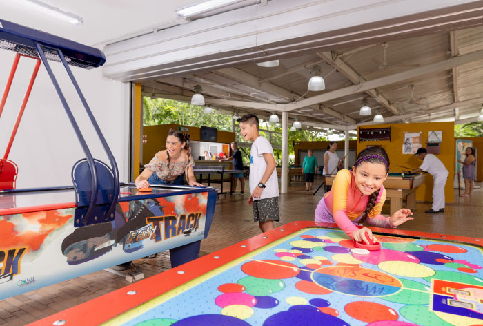 Tres personas que están jugando en mesas de hockey, dentro de un salón de juegos que tiene paredes de color mostaza y tejas blancas, luego de adquirir uno de los planes vacacionales en Lagosol