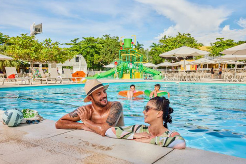 Un hombre y una mujer compartiendo dentro de una gran piscina sonríen y disfrutan uno de los  planes en el hotel Lagosol  para compartir en familia. Al fondo se ven dos niños con flotadores, un tobogán de color verde y carpas y sillas de color blanco