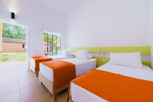 Habitación con descuentos especiales del centro vacacional Las Palmeras, que cuenta con tres camas personales con tendidos naranjas y blancos, cercanas a la puerta de salida que conecta con un prado