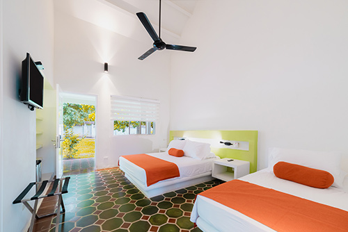 Habitación con baldosas de color verde oliva y moradas en donde se encuentran dos camas dobles con tendidos blancos y naranjas, un televisor, mesa de noche, ventilador y una puerta que da salida a un jardín del hotel Las Palmeras