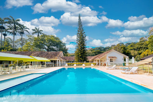 Piscina de 100 metros del hotel Las Palmeras con sillas playeras y mesas cubiertas con sombrillas, igual que palmeras, pinos y demás árboles alrededor del patio de una casa