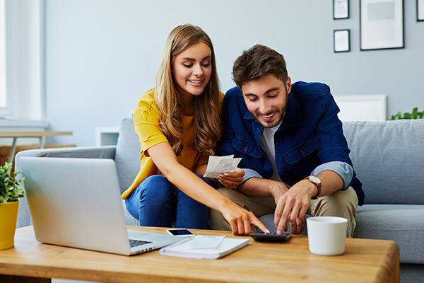 Pareja sentada en el sofá de la sala con un computador y una calculadora que están en una mesa de madera, revisando los pasos para diligenciar los métodos de pago de los planes de hoteles Compensar