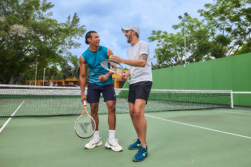 Dos hombres que usan ropa deportiva están hablando en medio de una cancha de tenis que tiene un piso y paredes de color verde a la cual se puede acceder luego de hacer una reserva de habitación en el hotel Lagosol