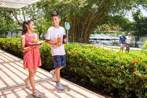 Dos niños, que usan ropa cómoda y llevan comida en sus manos, están caminando por un sendero rodeado de naturaleza y de un lago ubicado dentro de las instalaciones del hotel y parque acuático Lagosol