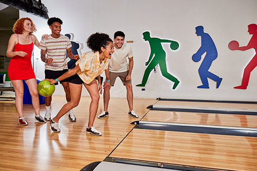 Dos hombres y dos mujeres en una bolera con siluetas de personas de colores verde, azul y rojo, en medio de una pared blanca, sonríen de emoción mientras disfrutan del plan de fin de semana festivo en el hotel Lagomar. La pista tiene piso de madera y se ven tres carriles de juego