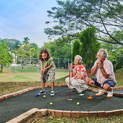 Dos adultos mayores compartiendo con un niño una partida de golf, en una cancha de color negro del alojamiento en Lagomar fin de semana festivo. El  entorno rodeado de una cancha sintética de fútbol y de naturaleza