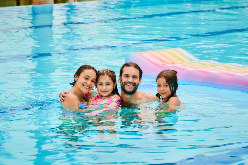 Una familia de un hombre, una mujer, un niño y una niña, están dentro de una piscina que tiene un colchón inflable de colores, flotando junto a ellos mientras sonríen, se relajan y aprovechan de las ofertas exclusivas para temporada alta en Lagomar