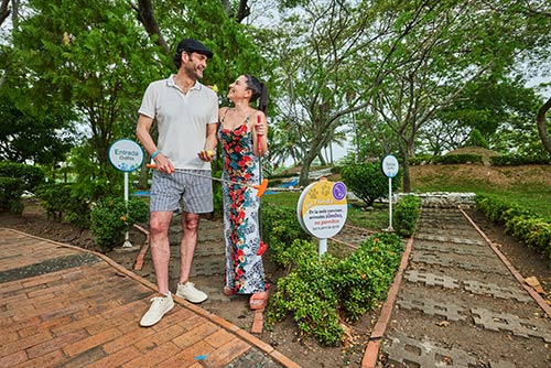 Pareja de un hombre vestido de camiseta gris, short gris de líneas y mujer con vestido de flores rojas y azules, se miran fijamente mientras sonríen, rodeados de naturaleza al enterarse de las tarifas del hotel Lagomar fin de semana y festivos