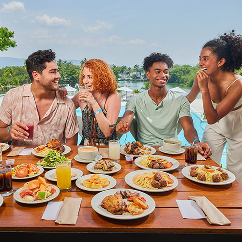 Dos hombres y dos mujeres, que visten de colores rosado claro, negro, verde menta y blanco, se encuentran sosteniendo vasos con bebidas y probando algunas preparaciones de comida que están en 18 platos sobre una mesa de madera rectangular en el Hotel Lagomar