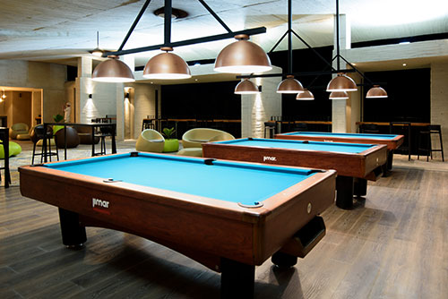 Sala de estar con muebles, puffs blancos y cafés, y lámparas que alumbran dos mesas de pool