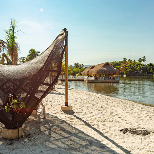 Parasol de madera con poli sombra de color negro y sillas blancas, detrás se puede visualizar una variedad de árboles y un quiosco con techo de hojas de palma y es uno de los beneficios de alquilar una habitación por una noche en Lagomar