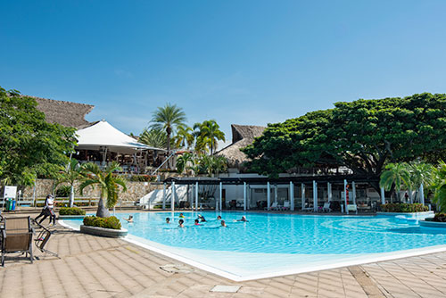 Piscina rodeada de palmeras, con personas disfrutando en el agua en un día soleado, luego de acceder al plan de alquiler de una habitación por una noche en Lagomar. Rodeada de gran cantidad de vegetación y acompañada de un cielo azul