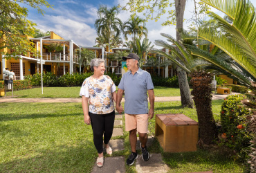 Pareja de personas mayores está caminando de la mano por un sendero que hace parte de los planes de hospedaje en el hotel Lagosol, el cual cuenta con habitaciones de paredes de color mostaza que están rodeadas de abundante naturaleza