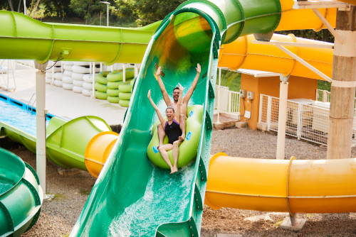Una mujer y un hombre que están descendiendo por un tobogán de colores verde y amarillo, levantan los brazos y sonríen luego de adquirir uno de los mejores planes vacacionales en Lagosol