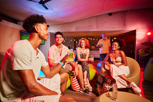 Grupo de amigos compartiendo un cóctel en uno de los espacios con ambientes neón incluidos con las ofertas de hoteles Compensar durante la temporada alta. Alrededor se ven más personas, una planta, un ventilador en el techo y al fondo un gran ventanal con una extensión de luces cálidas