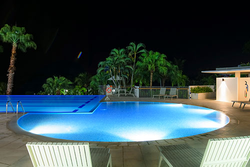 Piscina iluminada en medio de la noche, rodeada de naturaleza y de las habitaciones del hotel Lagomar en temporada alta. En el entorno se ven sillas asoleadoras y una construcción de paredes blancas