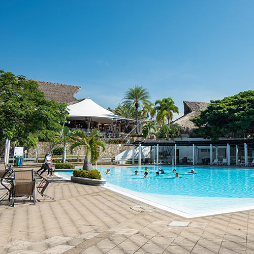 Gran piscina que tiene diez personas adentro, en medio de un día soleado y perfecto para aprovechar las ofertas de temporada alta Lagomar. Alrededor se ven sillas asoleadoras, un gran parasol blanco y dos grandes techos de paja
