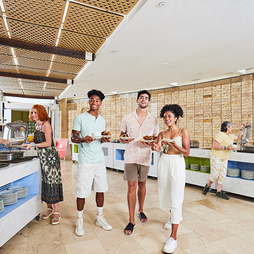 Grupo de amigos compuesto por dos hombres y dos mujeres, sostienen los platos de comida durante las mejores vacaciones en el hotel Lagomar temporada alta. Alrededor hay un ambiente de buffet, donde se ven platos y un espacio amplio