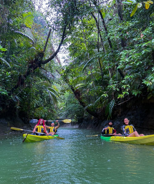 Cuatro personas en dos kayaks, uno amarillo y otro verde, reman por un río de aguas tranquilas rodeado de vegetación. Las personas usan chalecos salvavidas y disfrutan de unas vacaciones en Isla Fuerte con deportes acuáticos, en medio de la naturaleza