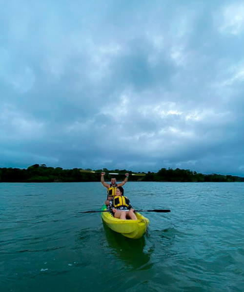 Dos personas, una de ellas con el remo encima de su cabeza y los brazos extendidos en modo de celebración; están navegando en un kayak de color amarillo luego de acceder a uno de los paquetes de viaje Isla Fuerte con ruta en kayak