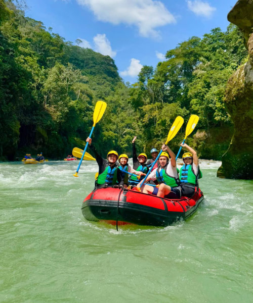 Grupo de turistas navegando en un bote inflable por un río de aguas verdes, usando chalecos salvavidas y remos amarillos, rodeados de vegetación y rocas naturales