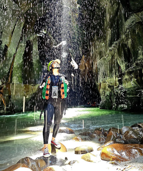 Mujer con casco y chaleco de seguridad caminando sobre rocas dentro de un cañón, mientras una cascada cae desde lo alto e ilumina el entorno rocoso