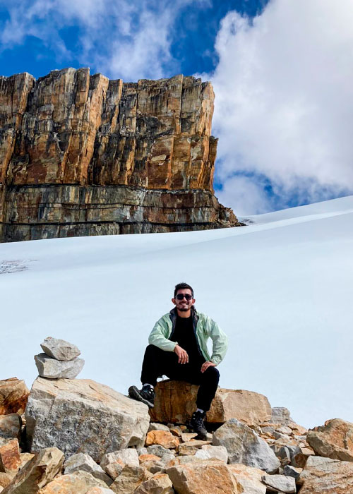 Hombre, que está sentado sobre una roca, sonríe y disfruta de uno de los planes de senderismo en la Sierra Nevada del Cocuy Colombia. En el fondo se ve la roca llamada el Pulpito del Diablo