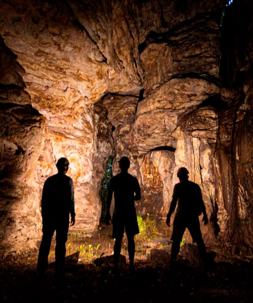 Tres personas, iluminando con linternas de luz cálida durante la exploración a una cueva que tiene formaciones rocosas, a la cual accedieron luego de comprar el viaje al Guaviare con guía incluido